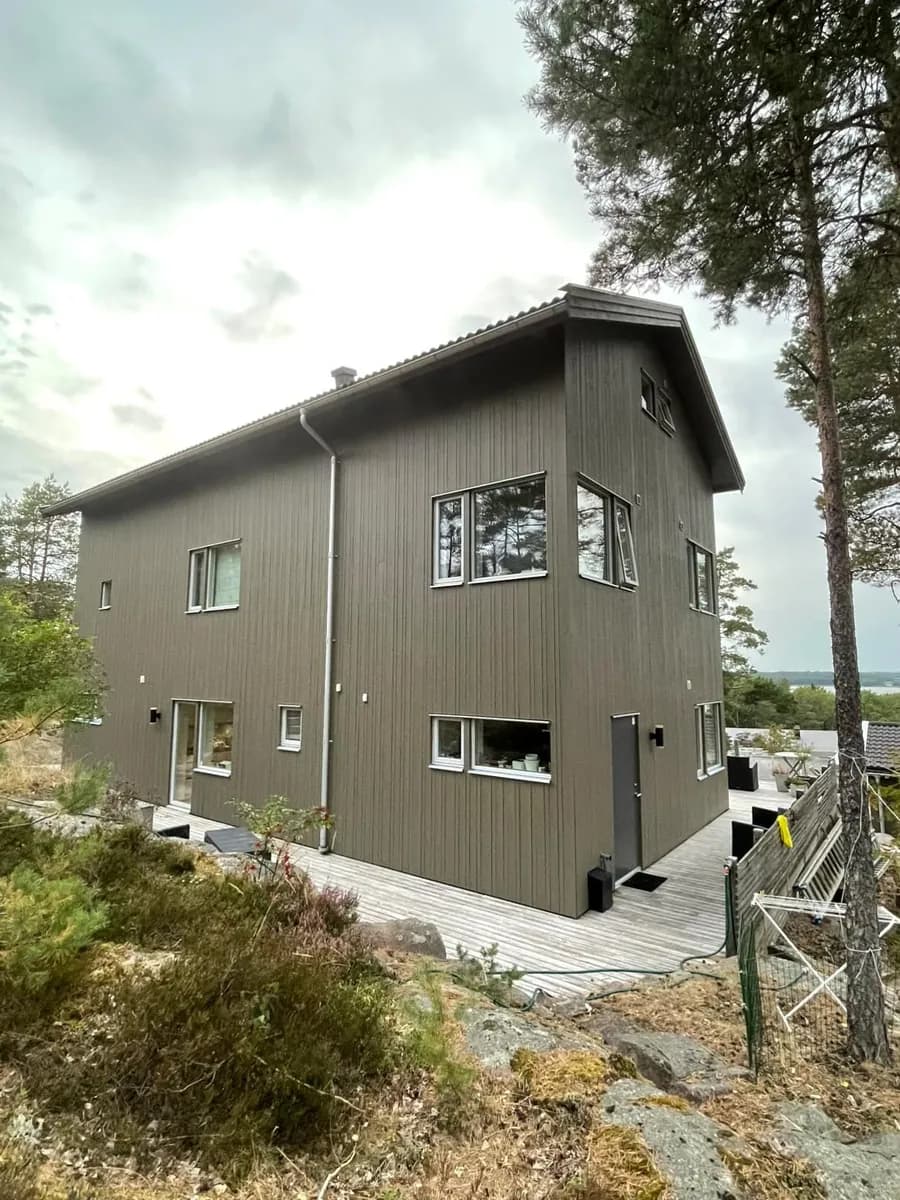 Samma nymålade grå träfasad i Täby kyrkby, sedd från tomten med tallar och berghällar