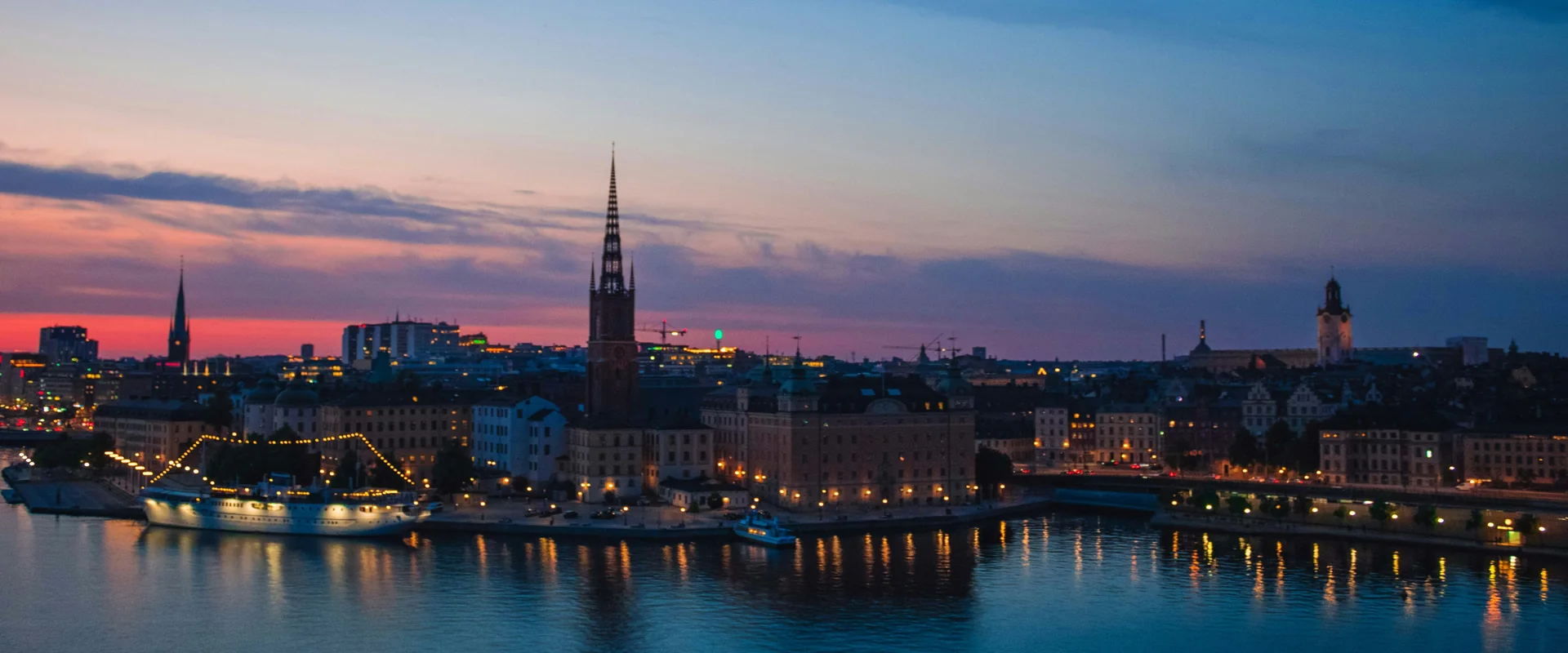 Stockholms siluett i skymning: Riddarholmskyrkans spira och Gamla stans fasader med tända fönster, speglade i Riddarfjärden.